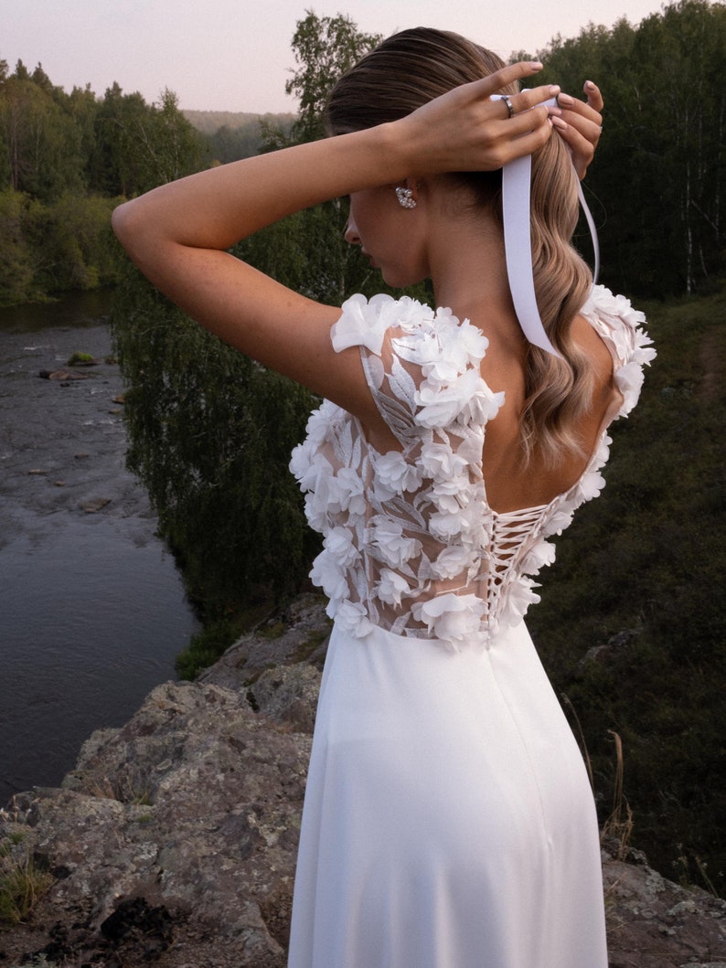 K&ouml;nnte beinhalten: Ein wei&szlig;es Brautkleid mit floralen Applikationen an Schultern und Oberteil. Das Kleid hat einen Schn&uuml;rr&uuml;cken und einen flie&szlig;enden Rock. Die Braut justiert ein wei&szlig;es Band in ihrem Haar, mit einem Fluss und B&auml;umen im Hintergrund.