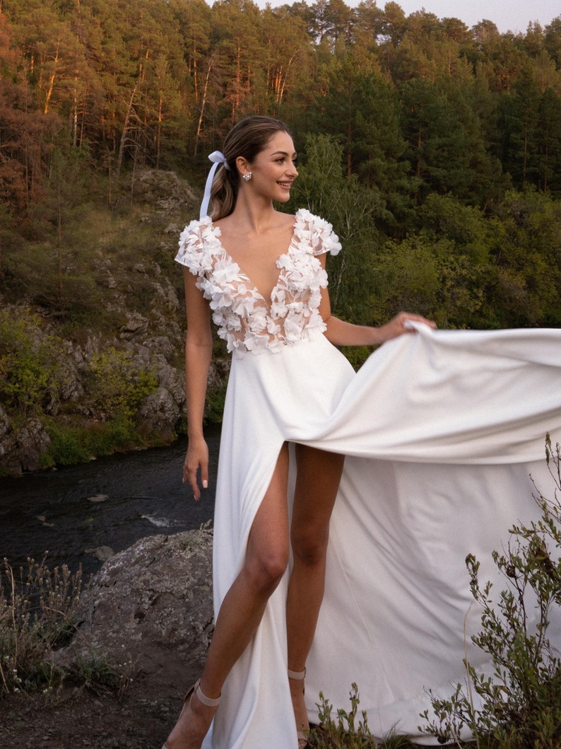 K&ouml;nnte beinhalten: Ein wei&szlig;es Hochzeitskleid mit tiefem V-Ausschnitt und floralen Verzierungen. Das Kleid hat einen hohen Schlitz und einen flie&szlig;enden Rock. Die Frau steht drau&szlig;en in der N&auml;he eines Flusses, mit einem Wald im Hintergrund.