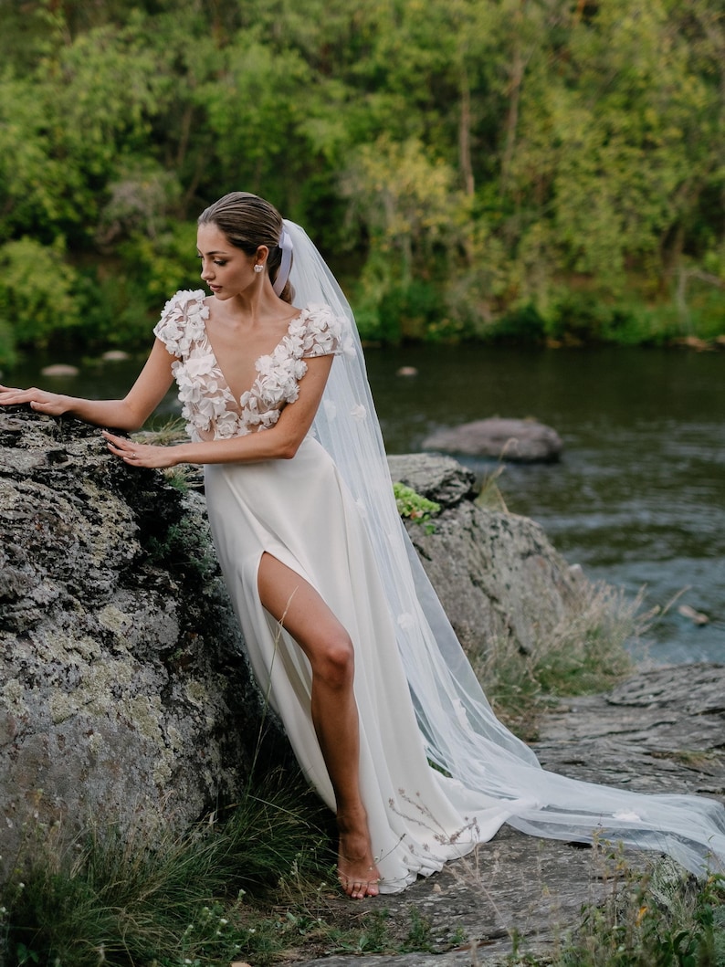 K&ouml;nnte beinhalten: Wei&szlig;es Hochzeitskleid mit hohem Schlitz und floralem Oberteil. Das Kleid hat einen langen, flie&szlig;enden Schleier mit zarten floralen Akzenten. Die Braut steht auf einem Felsen in der N&auml;he eines Flusses, mit einem nat&uuml;rlichen gr&uuml;nen Hintergrund.