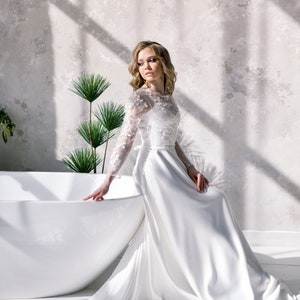 May include: A white wedding dress with floral lace sleeves and bodice, featuring a flowing skirt. The bride is seated near a white bathtub, with sunlight casting shadows on the textured wall behind her.