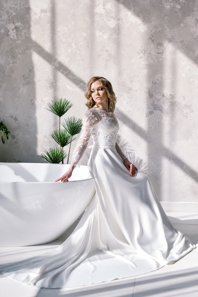 May include: A white wedding dress with floral lace sleeves and bodice, featuring a flowing skirt. The bride is seated near a white bathtub, with sunlight casting shadows on the textured wall behind her.