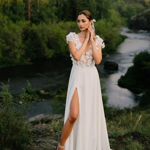 K&ouml;nnte beinhalten: Wei&szlig;es Hochzeitskleid mit hohem Schlitz und floralem Oberteil. Das Kleid wird im Freien mit einem Fluss und B&auml;umen im Hintergrund getragen. Das Kleid hat einen flie&szlig;enden Rock und wird mit neutralfarbenen Abs&auml;tzen kombiniert.