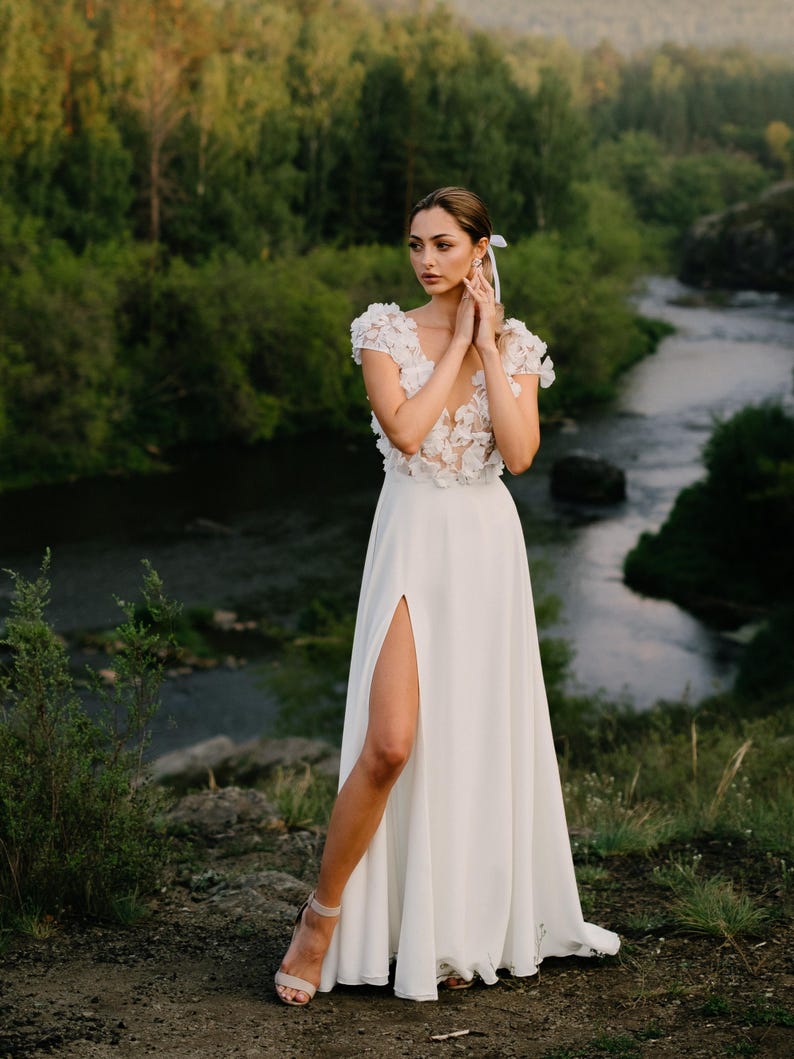 K&ouml;nnte beinhalten: Wei&szlig;es Hochzeitskleid mit hohem Schlitz und floralem Oberteil. Das Kleid wird im Freien mit einem Fluss und B&auml;umen im Hintergrund getragen. Das Kleid hat einen flie&szlig;enden Rock und wird mit neutralfarbenen Abs&auml;tzen kombiniert.