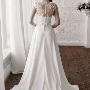May include: A white wedding dress with a long train. The bodice features floral embroidery on the sheer sleeves and back. The dress has a button-up back and a small bow at the waist. The bride is standing in front of a window.