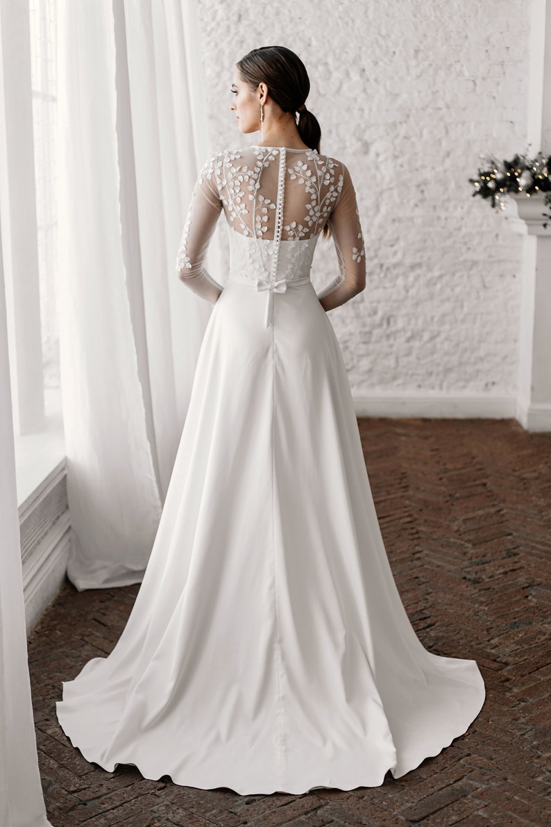 May include: A white wedding dress with a long train. The bodice features floral embroidery on the sheer sleeves and back. The dress has a button-up back and a small bow at the waist. The bride is standing in front of a window.