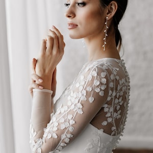 May include: A bride in a white wedding dress with long sleeves and floral lace detailing. The dress features a row of buttons down the back. The bride is wearing long, delicate earrings with a leaf design.
