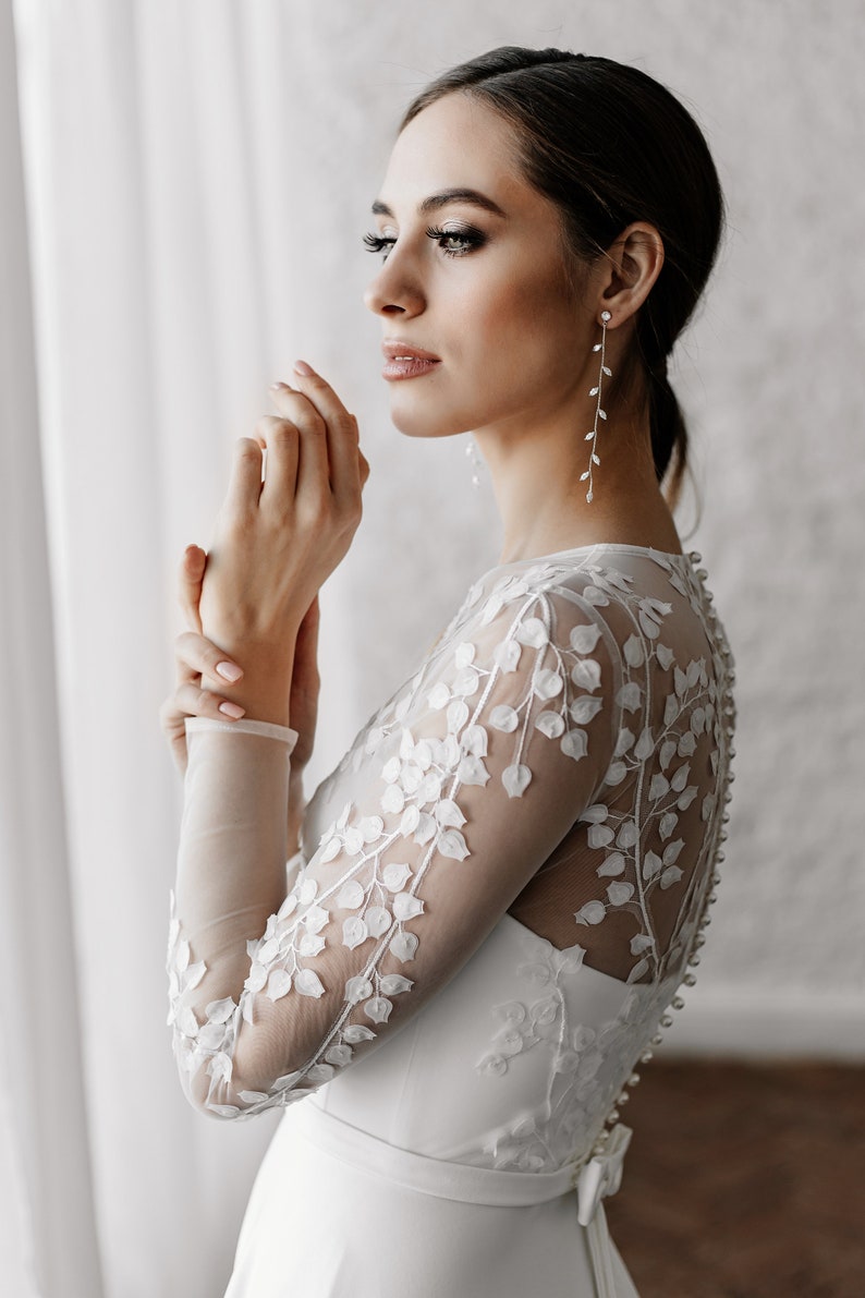 May include: A bride in a white wedding dress with long sleeves and floral lace detailing. The dress features a row of buttons down the back. The bride is wearing long, delicate earrings with a leaf design.