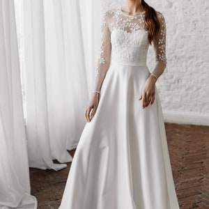May include: A white wedding dress with a floral embroidered bodice and sheer long sleeves. The dress has a full, flowing skirt and a simple, elegant design. The bride is wearing a bracelet and earrings.