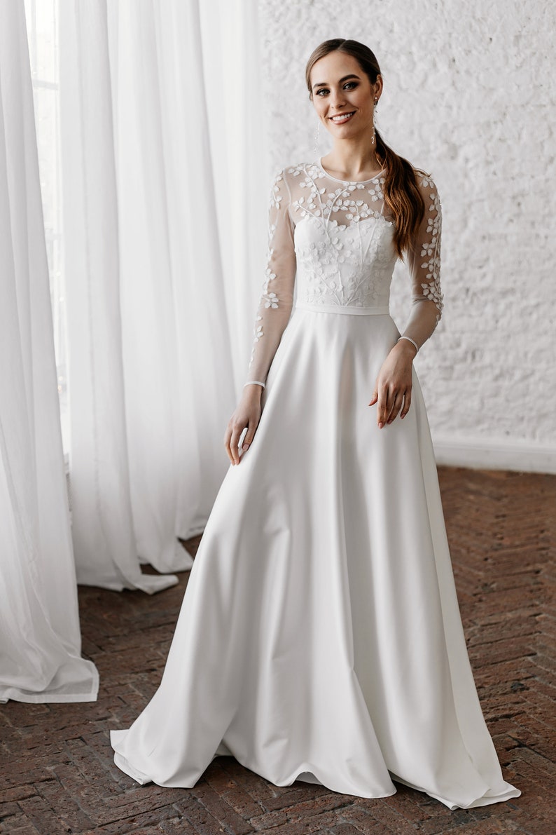 May include: A white wedding dress with a floral embroidered bodice and sheer long sleeves. The dress has a full, flowing skirt and a simple, elegant design. The bride is wearing a bracelet and earrings.