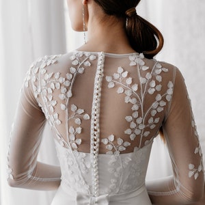 May include: A white wedding dress featuring a sheer back with delicate floral embroidery. The dress has long sleeves, a row of buttons down the back, and a bow at the waist. The overall design is elegant and romantic.