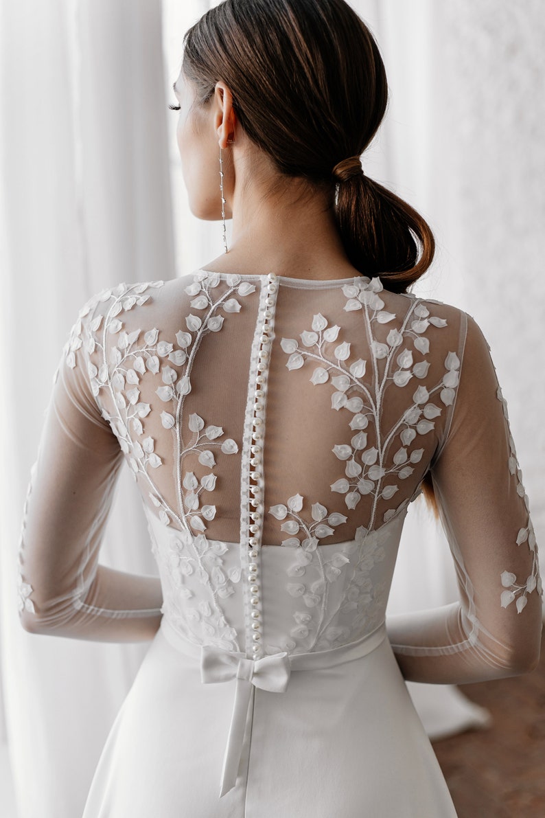 May include: A white wedding dress featuring a sheer back with delicate floral embroidery. The dress has long sleeves, a row of buttons down the back, and a bow at the waist. The overall design is elegant and romantic.