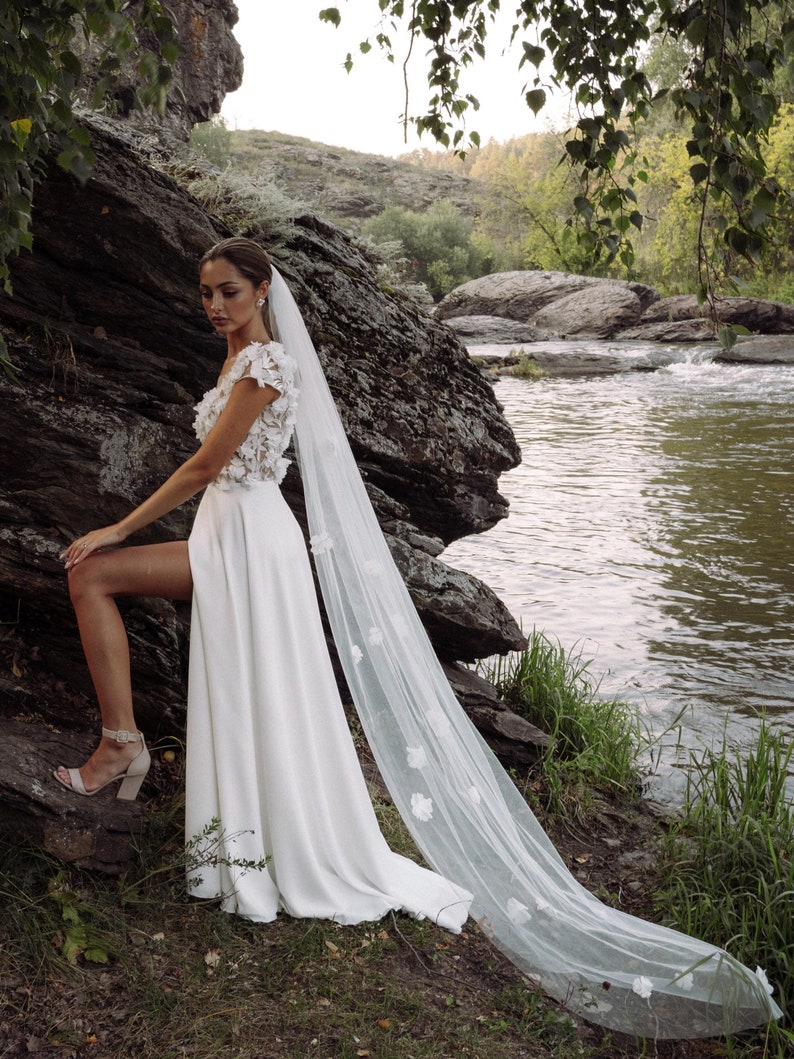 K&ouml;nnte beinhalten: Wei&szlig;es Hochzeitskleid mit hohem Schlitz und floralem Oberteil. Das Kleid hat einen langen, flie&szlig;enden Schleier, der mit zarten floralen Applikationen verziert ist. Die Braut tr&auml;gt beige Abs&auml;tze und posiert in der N&auml;he eines Flusses.