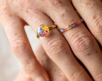 May include: Two gold rings, one with a clear, round setting filled with colorful dried flowers, and the other a thin band with a floral pattern. The rings are on a person's finger, showcasing the jewelry's design and details.