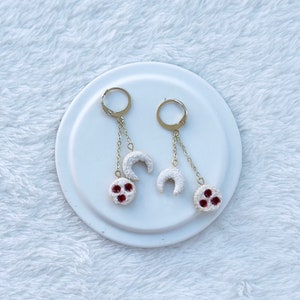Könnte beinhalten: Ein Paar goldfarbene Hängeohrringe. Jeder Ohrring hat einen goldenen Reifen, eine Kette, einen weißen Halbmond und einen weißen kreisförmigen Anhänger mit roten Akzenten. Die Ohrringe werden auf einem weißen Teller vor einem weißen, strukturierten Hintergrund präsentiert.