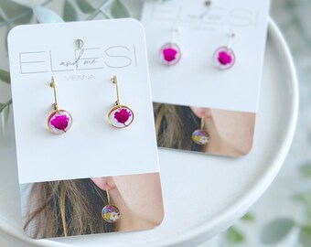 May include: Two pairs of dangle earrings on white cards. The earrings feature a small, round, gold-colored frame with a vibrant pink flower encased in clear resin. The cards have the text "ELLES and me VIENNA".