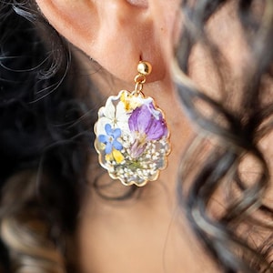 May include: Gold-colored dangle earring with a clear resin oval pendant. The pendant is filled with a variety of dried flowers in shades of purple, white, blue, and yellow. The earring is attached to the ear with a small gold ball.