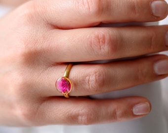 May include: A gold ring with a vibrant pink, oval-shaped stone. The ring is worn on a finger, with the hand resting against a white shirt. The ring's band has a simple, elegant design.