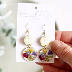 Peut inclure: Boucles d'oreilles en résine transparente et dorée avec des fleurs pressées. Les boucles d'oreilles présentent un disque rond doré et un disque rond en résine transparente avec une variété de fleurs pressées dans des tons de rouge, jaune, violet et bleu.