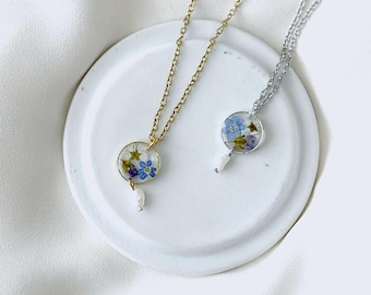 May include: Two delicate necklaces with floral pendants. One necklace is gold-toned, the other silver-toned. Each pendant features a clear, circular resin encasing small blue, purple, and white flowers. A small, white pearl hangs from each pendant.