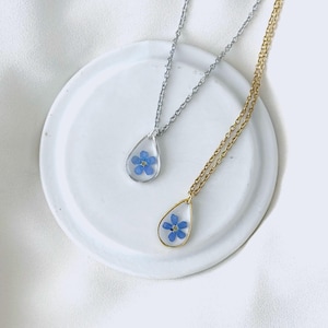 May include: Two teardrop-shaped pendant necklaces. One has a silver chain and frame, the other a gold chain and frame. Each pendant contains a small blue flower. The necklaces rest on a white, round, ceramic dish.