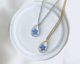 May include: Two teardrop-shaped pendant necklaces. One has a silver chain and frame, the other a gold chain and frame. Each pendant contains a small blue flower. The necklaces rest on a white, round, ceramic dish.