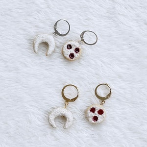 Puede incluir: Un par de pendientes de plata y oro con forma de media luna y una galleta redonda con relleno rojo. Los pendientes están sobre una superficie blanca y peluda.