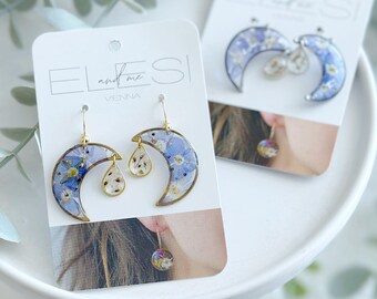 May include: Two pairs of earrings on white cards. The earrings are crescent moon-shaped, filled with blue and white flowers, and have gold-colored metal accents. One pair has teardrop-shaped dangles, and the other has a small round dangle.