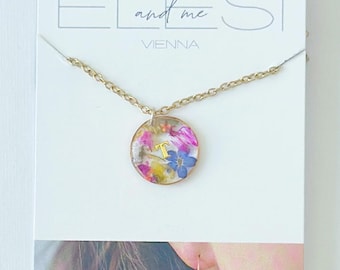 May include: A gold chain necklace with a circular pendant containing pressed colorful flowers and a gold letter T. The jewelry is displayed on a white card labeled EL and me VIENNA, which also shows a matching circular earring with pressed flowers.