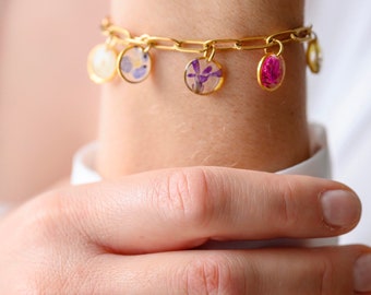 May include: Gold-toned bracelet with multiple small, round charms containing pressed flowers in shades of purple, pink, and white. A matching gold ring with a pink flower is also visible. The jewelry is worn on a wrist.