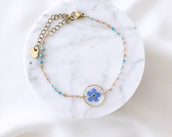 May include: A delicate gold bracelet with a small blue flower encased in resin. The bracelet features a heart-shaped clasp and is adorned with tiny colorful beads.