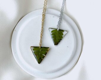 May include: Two necklaces with triangular pendants, each featuring a green leaf encased in clear resin. One necklace has a gold chain and the other a silver chain. The pendants are displayed on a white, circular dish.