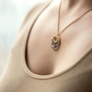 Könnte beinhalten: Eine goldfarbene Halskette mit einem Anhänger mit getrockneten Blumen. Der ovale Anhänger ist mit einer Mischung aus bunten getrockneten Blumen gefüllt und hängt an einer zarten Goldkette. Die Halskette wird von einer Person getragen.