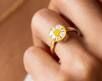May include: A gold ring featuring a daisy flower design. The ring has a round, yellow and white flower set in a gold band. The ring is shown on a person's finger.