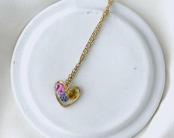 May include: A gold-colored necklace with a heart-shaped pendant. The pendant is filled with colorful dried flowers. The necklace is displayed on a white, round, ceramic plate. The chain is delicate and thin.