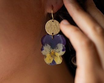 May include: Gold-colored earrings featuring a circular top and a pendant shaped like a pansy flower. The pansy pendant has purple and yellow petals. The earrings are hanging from a person's ear.
