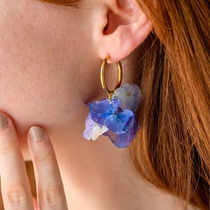 Könnte beinhalten: Goldene Creolen mit Büscheln aus getrockneten blauen und lila Blumen. Ein goldener Ring mit einem rosa Blumendesign ist ebenfalls zu sehen. Die Ohrringe und der Ring heben sich vor dem Hintergrund von rotem Haar und heller Haut ab.