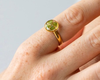 May include: A gold-colored ring with a round, clear setting. The setting contains a green, plant-like design. The ring is on a finger, showcasing the jewelry's design and color.