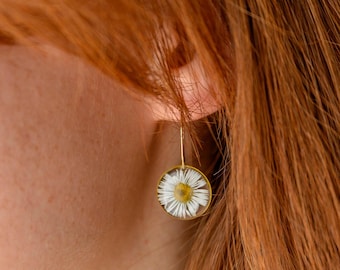 May include: Gold-tone dangle earrings featuring a preserved daisy flower encased in a clear, round resin. The daisy has white petals and a yellow center. The earrings are shown against a background of red hair and skin.