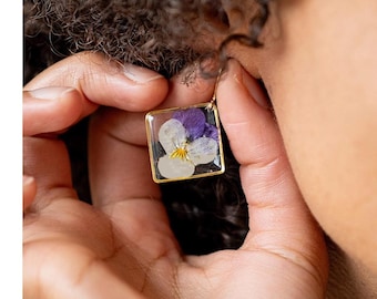 May include: A square gold-toned earring with a pressed flower design. The flower has white, purple, and yellow hues, encased in clear resin. The earring is being held near an ear.