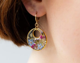May include: Gold-tone hoop earrings with a clear resin center filled with colorful dried flowers. The flowers include shades of blue, pink, and white. The earrings are round and have a delicate, feminine design.