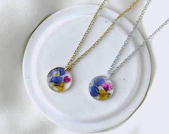 May include: Two delicate necklaces with round pendants, each showcasing a vibrant arrangement of dried flowers encased in clear resin. One necklace has a gold chain, the other a silver chain. The pendants feature blue, pink, and yellow floral elements.