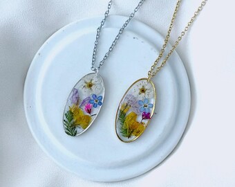 May include: Two oval pendant necklaces with dried flowers encased in clear resin. One necklace has a silver chain, the other a gold chain. The pendants feature yellow, purple, and blue flowers and green foliage. The necklaces rest on a white ceramic dish.