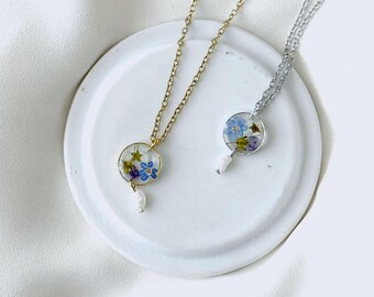 May include: Two delicate necklaces with floral pendants. One necklace has a gold chain and pendant, the other has a silver chain and pendant. Each pendant is a clear, circular resin containing small blue, purple, and white flowers, with a small white pearl drop.