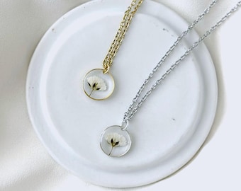 May include: Two necklaces with delicate white flowers encased in clear resin pendants. One necklace has a gold chain, the other a silver chain. Both pendants are round and sit on a white plate.