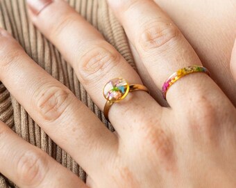 May include: Two gold rings with colorful floral designs. One ring features a round, clear resin setting with embedded flowers. The other ring is a thin band with a floral pattern. The rings are on a hand with a neutral background.