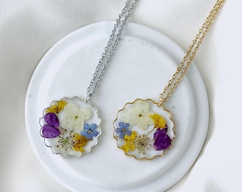 May include: Two floral pendant necklaces. One has a silver chain and bezel, the other a gold chain and bezel. Both pendants feature a clear resin with a variety of dried flowers in shades of yellow, purple, white, and blue.