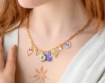 May include: A gold-colored chain necklace with various charms. Charms include a diamond shape with pink accents, a bee, a daisy, purple flowers, a yellow triangle, a silver disc, and a blue flower. The necklace is worn on a person.