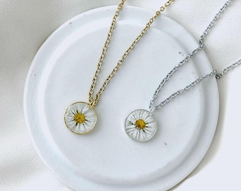 May include: Two daisy pendant necklaces. One necklace has a gold chain and bezel, the other has a silver chain and bezel. Each pendant features a white daisy with a yellow center encased in clear resin. The necklaces rest on a white, round dish.