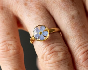 May include: A gold-colored ring featuring a clear, round setting encasing a delicate blue and yellow flower. The ring is on a finger, showcasing the floral design and the ring's polished finish.