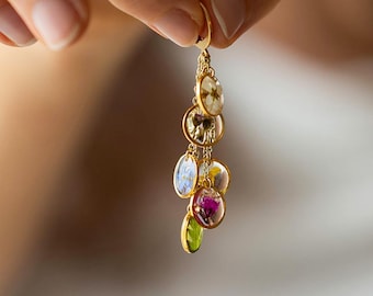 May include: A gold-toned earring featuring a cascade of oval pendants. Each pendant showcases a preserved flower in various colors, including white, blue, purple, and green. The earring is held by a hand, highlighting the delicate floral design.